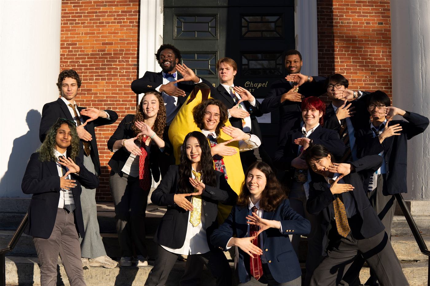 The Zumbyes on the chapel steps in blazers and ties, framing their faces with their hands. One member is wearing a full banana suit.